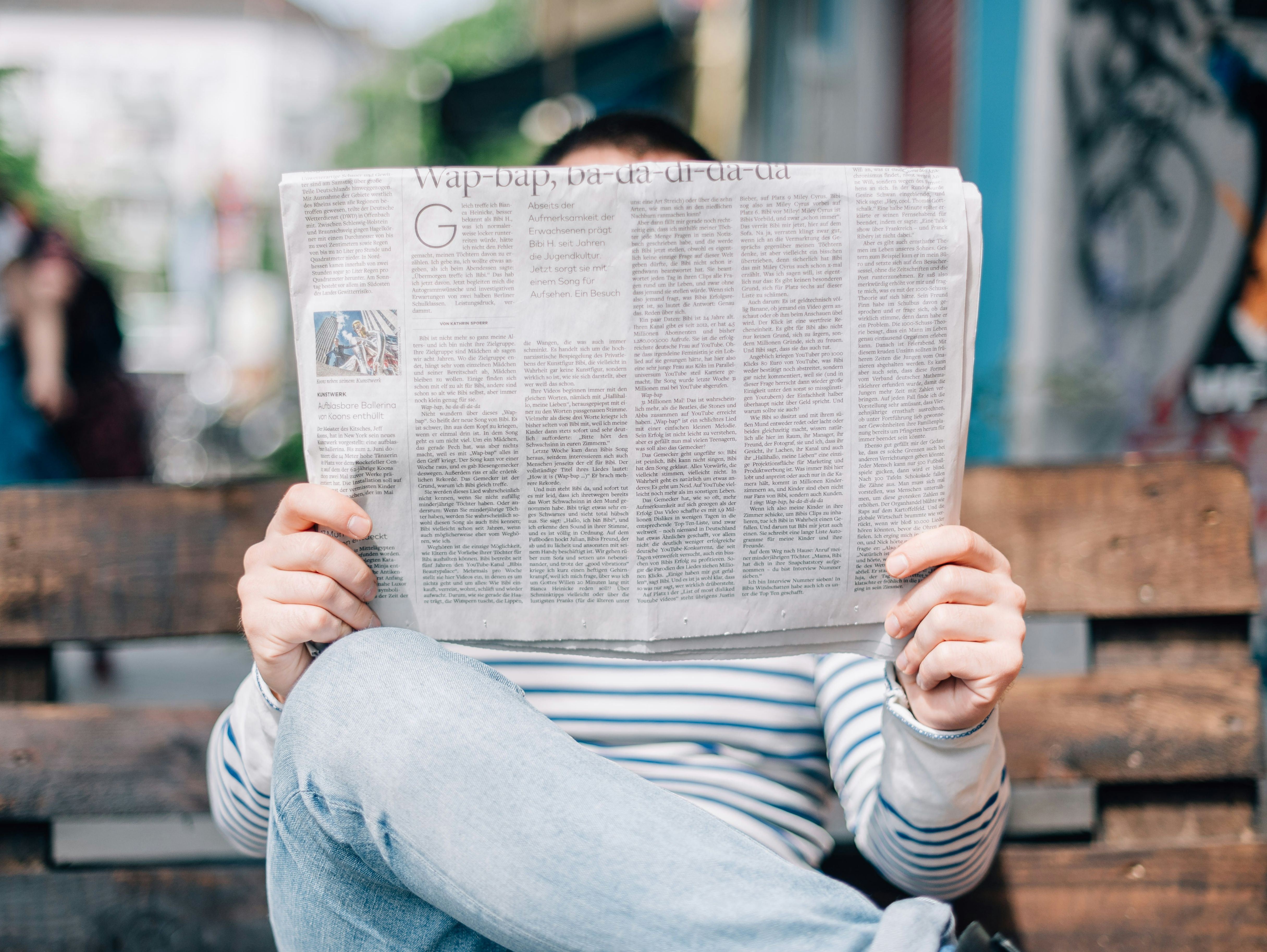 Person sitzt verdeckt von einer Zeitung auf einer Bank und liest